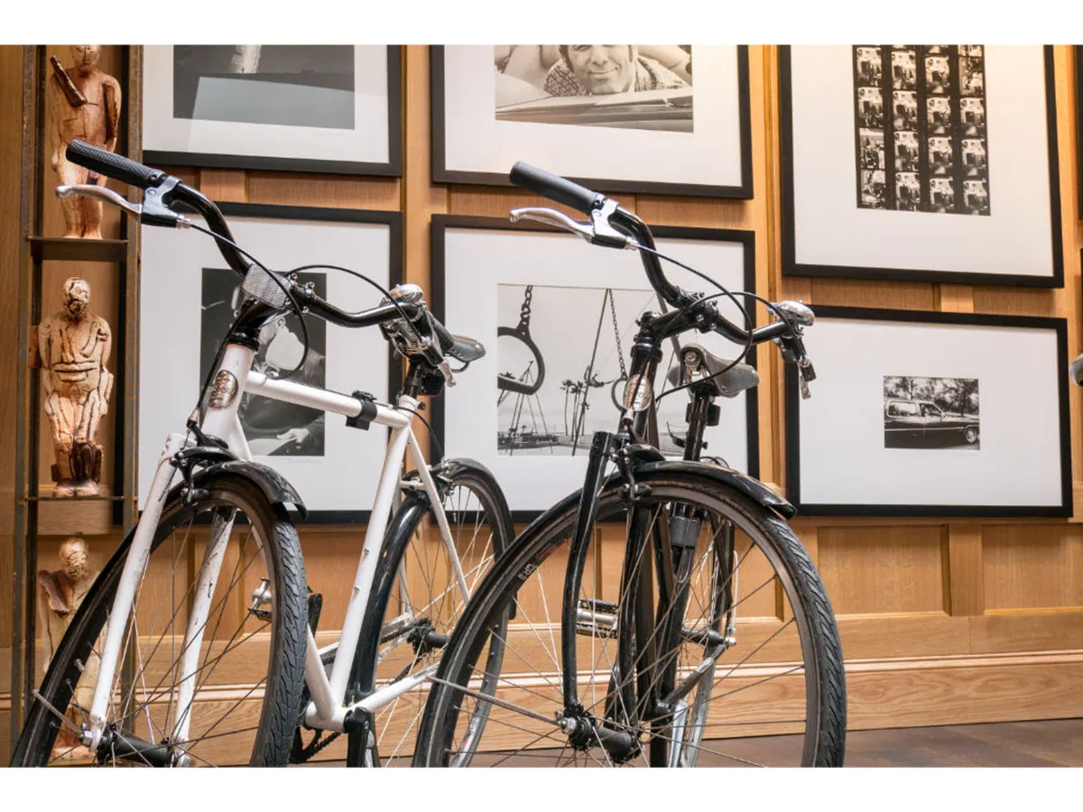 A creative lounge area featuring two stylish bicycles (one black, one white) displayed against a textured wooden wall, complemented by framed black and white art photographs. The setting evokes a modern, relaxed vibe ideal for guests interested in exploring the city via the hotel's Shinola bicycle rentals.