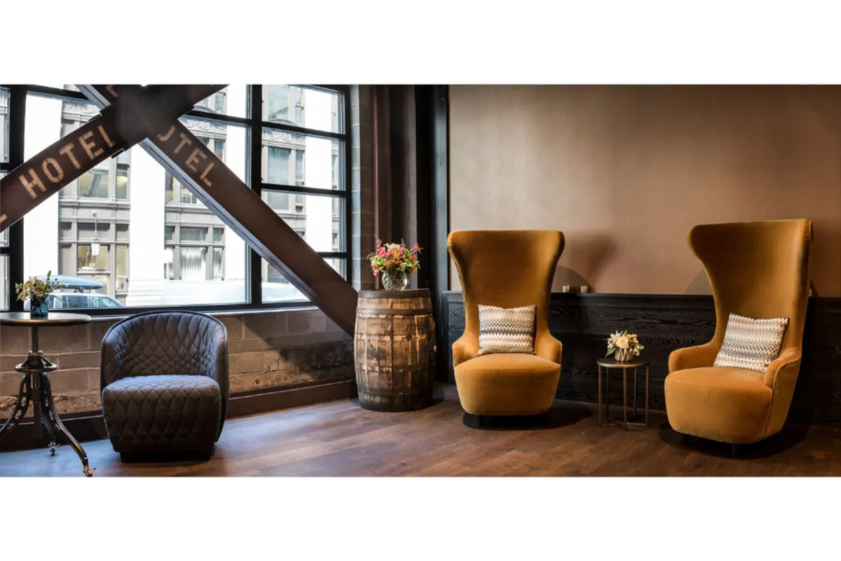 A cozy lounge area with two high-backed chairs, a wooden barrel serving as a side table, and large windows that let in natural light, creating an inviting connection to the outdoors. The seating area is perfect for relaxing after exploring the city, and the windows suggest an urban backdrop just outside.