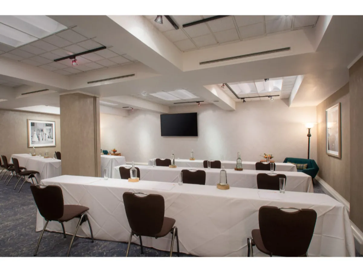A formal interior space featuring long tables with white tablecloths, a black presentation screen, and subtle decorative touches. The room is well-lit, designed for business meetings, conferences, workshops, and networking events.