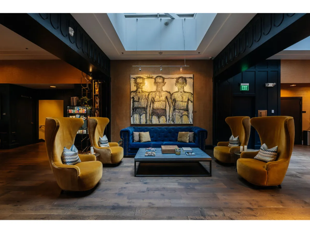 A vibrant public lounge with a blue sofa, yellow armchairs, artistic wall displays, and a central coffee table. The setting is ideal for socializing, relaxing with a book from the spiritual menu, or engaging in lively conversation. The creative decor supports the hotel's artistic and welcoming theme.