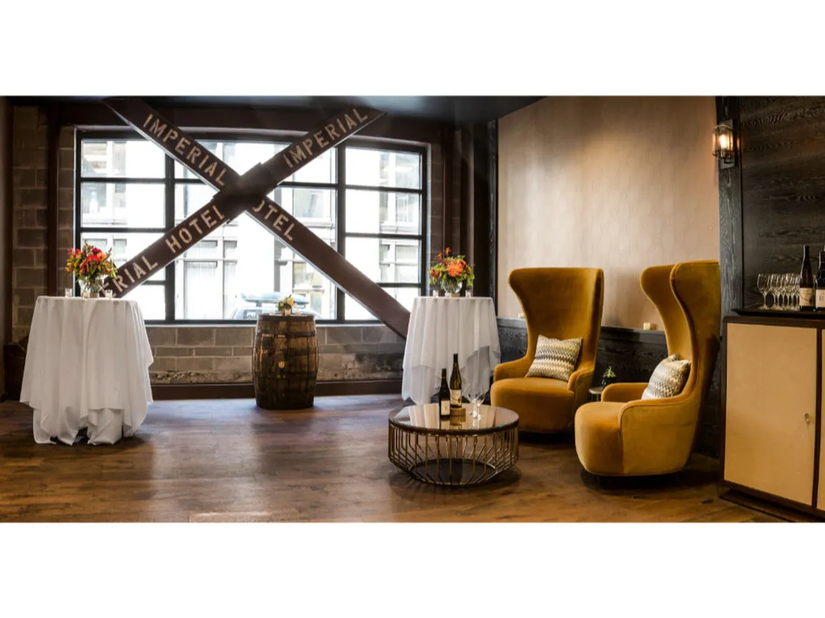 A stylish lounge area with golden armchairs, floral arrangements, barrel tables, and a window with the 'IMPERIAL HOTEL' sign. The scene conveys a warm, welcoming mood, ideal for intimate conversations, wine tastings, and celebratory gatherings after a busy workday.