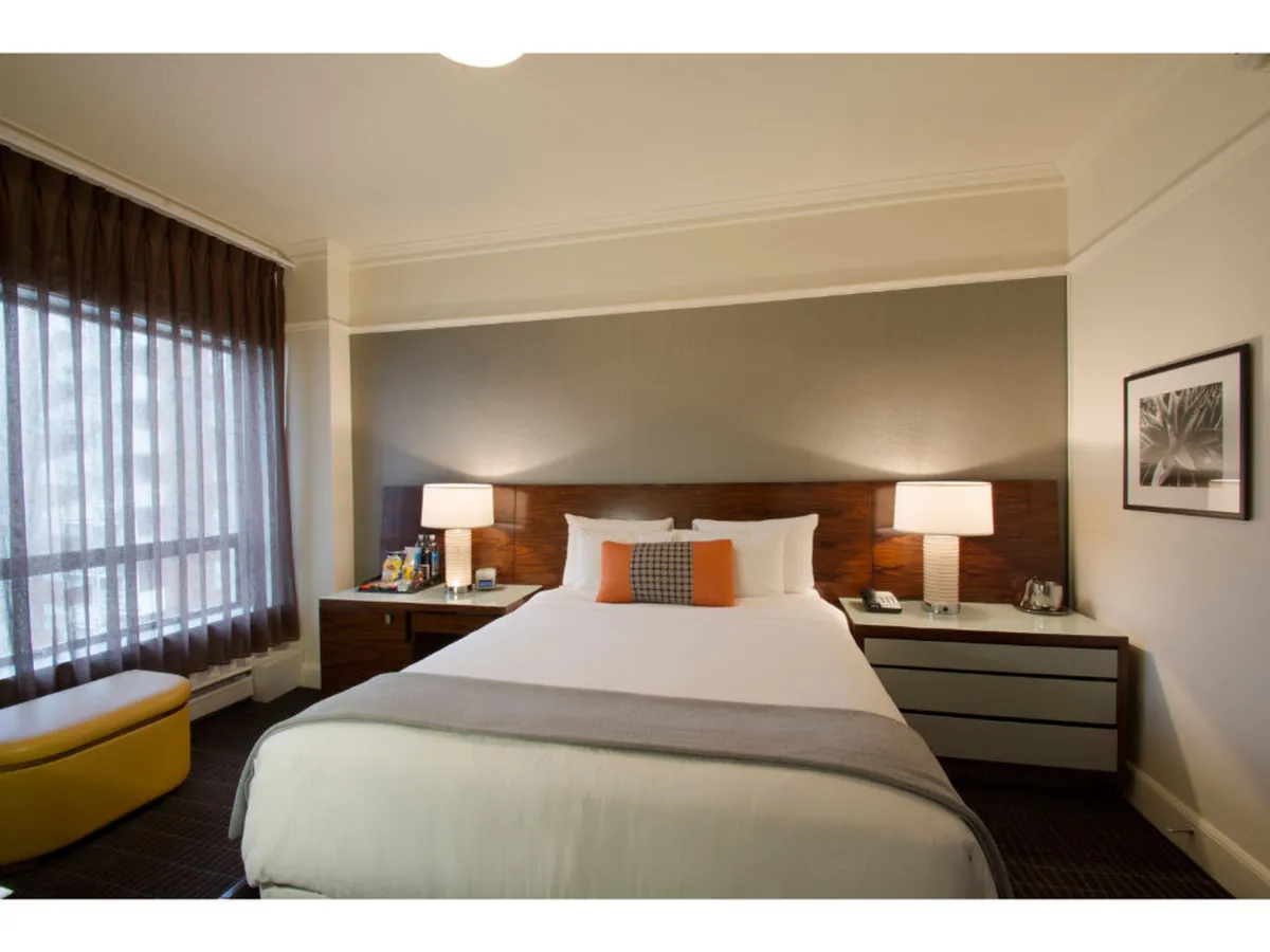 The image features a chic hotel room with a plush bed adorned with decorative pillows, flanked by stylish nightstands and warm lamps. Sunlight streams in through a large window with elegant curtains, creating a serene and inviting atmosphere. The scene suggests a perfect spot for relaxation, enjoying a book, or sipping a morning coffee while bathed in natural light.