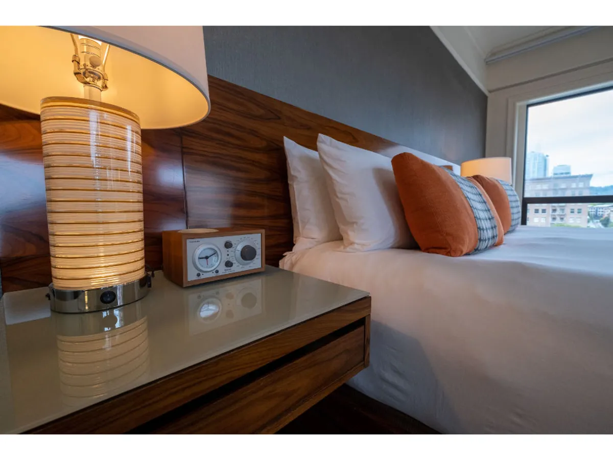A tranquil Premium Queen room featuring a plush queen-size bed adorned with decorative pillows, a soft lamp on the nightstand, and a clock radio. The cozy setting is suffused with natural morning light streaming in through the windows, creating a welcoming and restful atmosphere. A coffee cup on the nightstand suggests the possibility of enjoying a morning drink in bed.