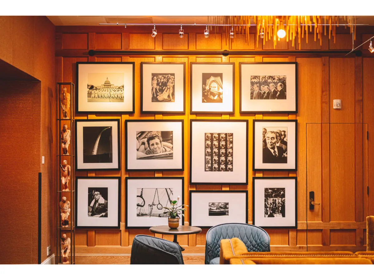 A warm, wood-paneled public space featuring a black and white photo gallery on the wall, a cozy seating area with a coffee table, and an indoor plant. The art display draws attention, suggesting the presence of the Kennerly photography collection, while the comfortable seating invites guests to linger and socialize.