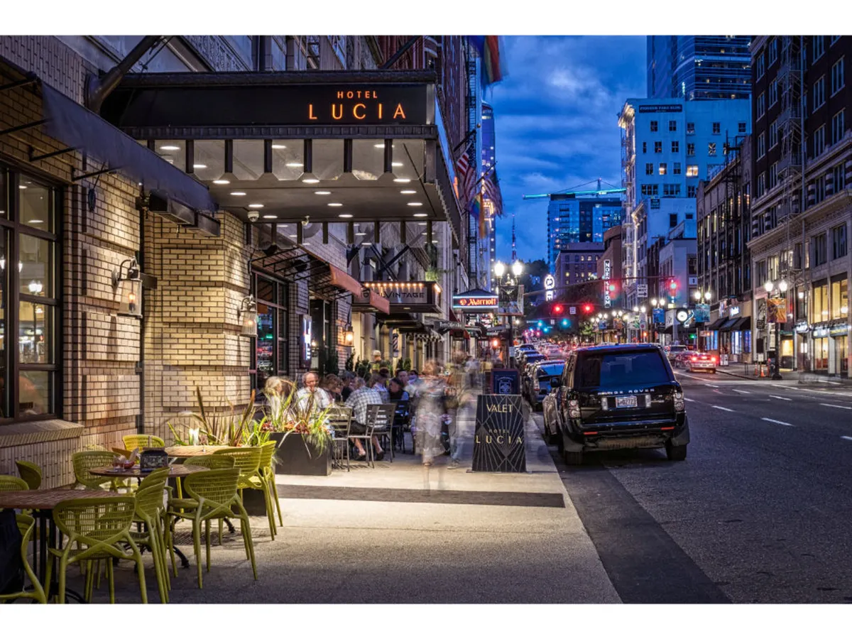 The image displays the exterior of Hotel Lucia, with its distinctive signage, outdoor seating area, vibrant street lights, and people enjoying the city ambiance. The lively sidewalk scene suggests a dynamic urban location, with easy access to downtown attractions and business venues.