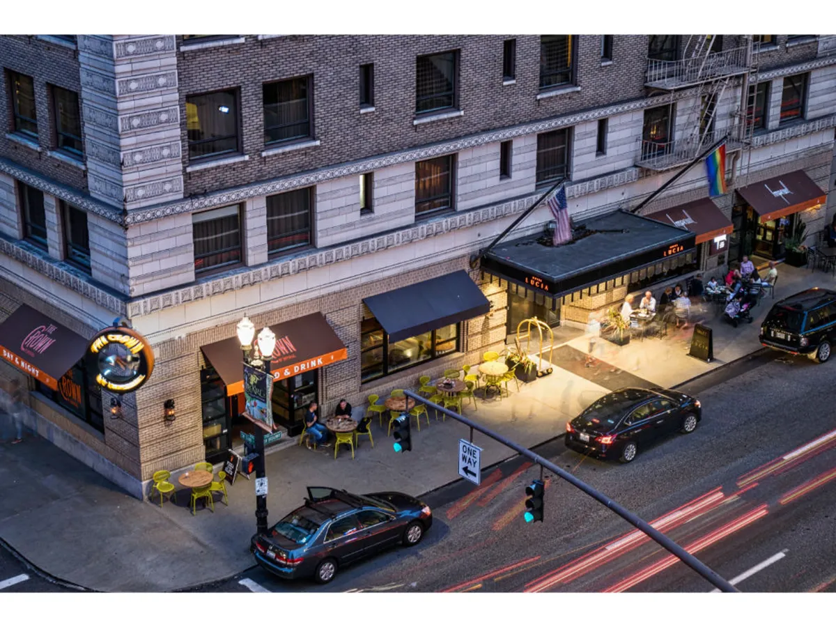 Depicts an inviting outdoor seating area with a restaurant sign and softly glowing street lights. The scene captures couples and friends dining al fresco, cars passing by, and a warm, sociable atmosphere suited to romantic evenings out.