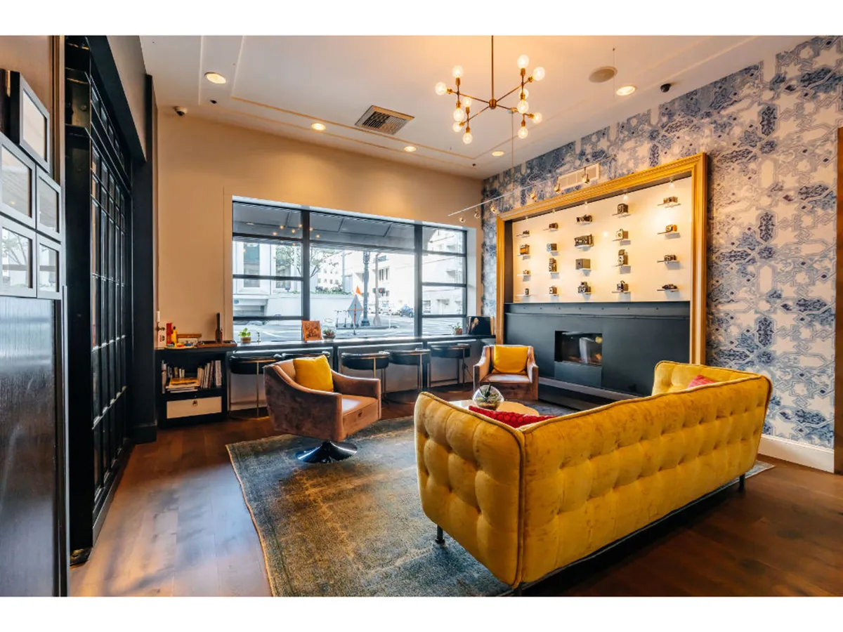 An inviting public lounge area with a yellow tufted sofa, elegant chandelier, decorative wall with shelves, and large windows letting in natural light. The setting is comfortable and trendy, ideal for relaxing, socializing, or catching up on work in a stylish public space.