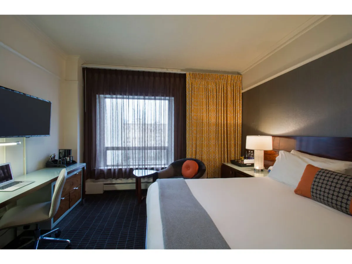 A serene guest room featuring a comfortable bed, a sunlit window with curtains, a functional desk and chair, and a tranquil atmosphere. The scene suggests a guest enjoying coffee by the window or unwinding with a book, highlighting comfort and practical amenities.